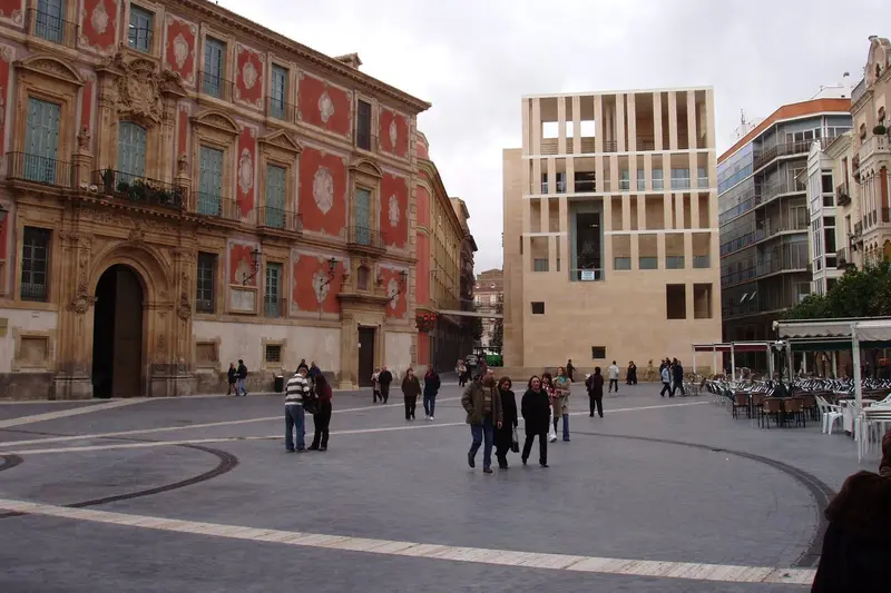 Paseantes cruzando la Plaza del Cardenal Belluga, frente al Palacio Episcopal y el edificio de Moneo, con terrazas a la derecha.
