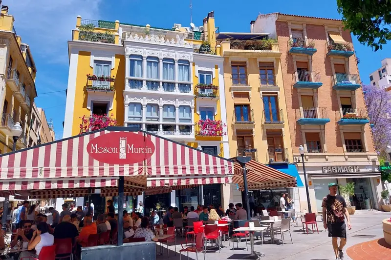 Terrazas llenas de gente bajo un toldo a rayas rojas y blancas en la Plaza de las Flores, con fachadas de colores y un hombre que cruza la plaza.