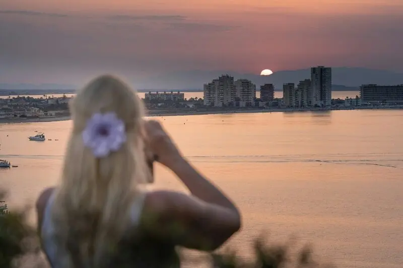 Una mujer de espaldas, con una flor en el pelo, fotografía la puesta de sol sobre el Mar Menor y los edificios de La Manga.