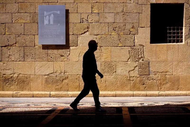 The silhouette of a man walking along the pavement beside a sunlit ashlar wall in the afternoon, in the centre of Murcia.